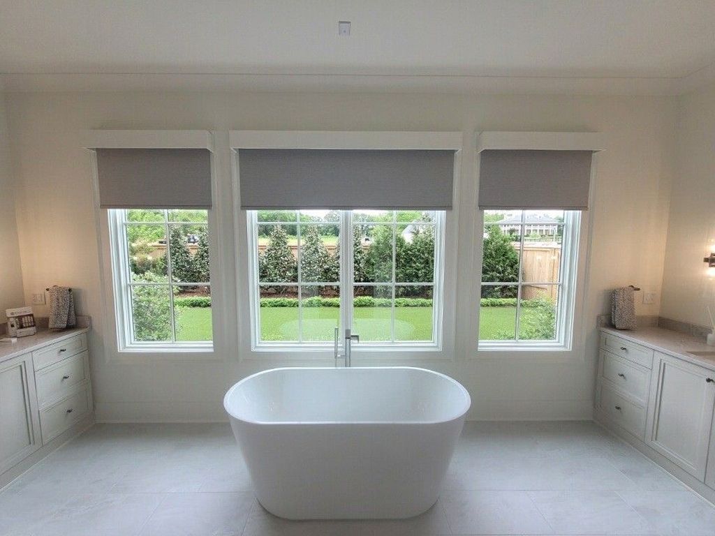 A luxurious bathroom with a freestanding white bathtub in front of three windows with grey shades, overlooking a green yard.