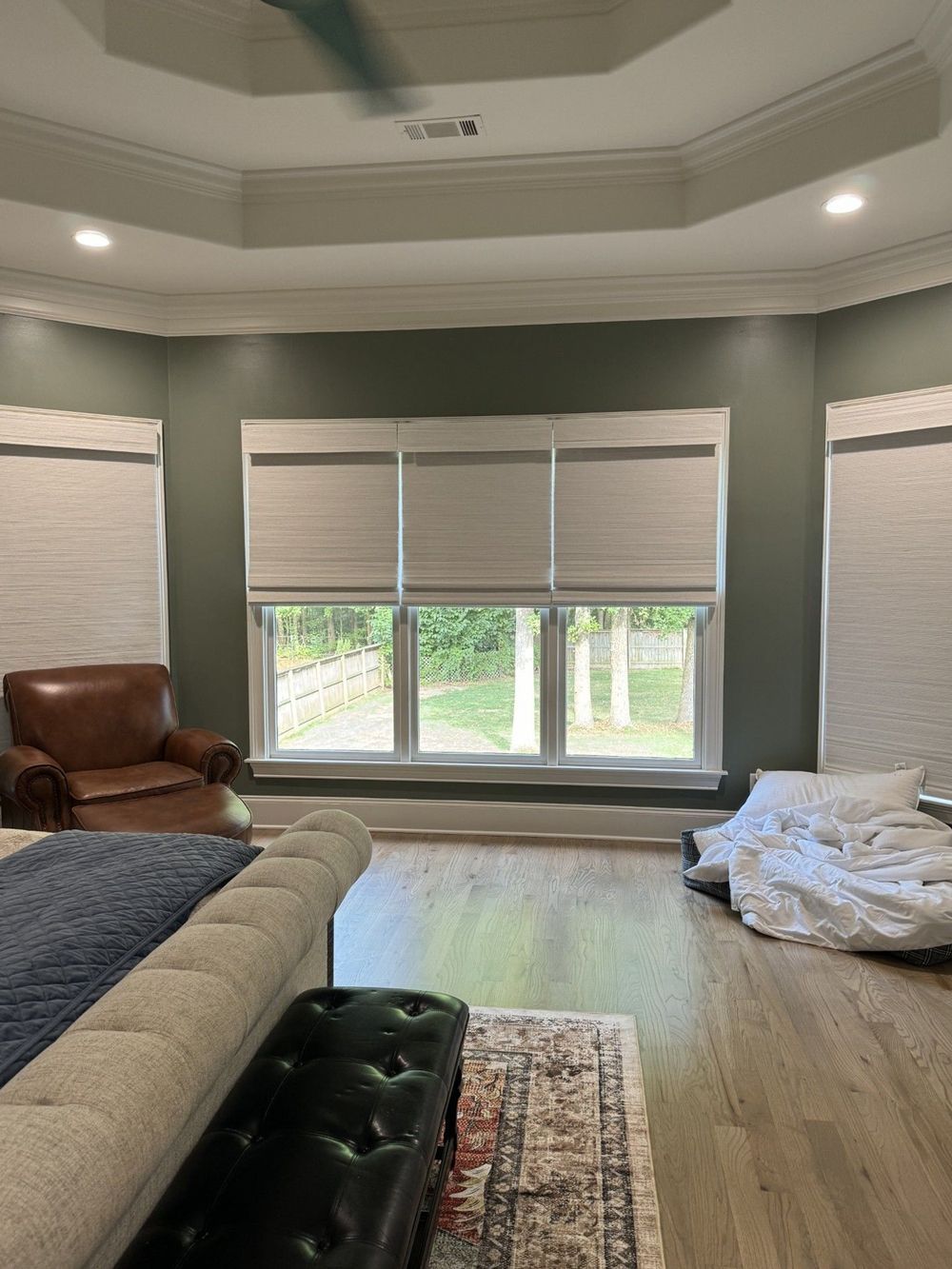 A bedroom with large windows and neutral-colored roman shades. An upholstered bed and seating area are visible in front of the windows.