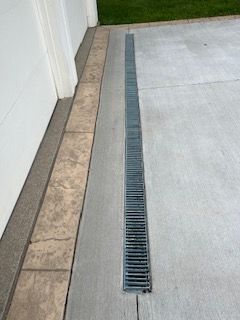 A drain on the side of a concrete driveway next to a garage door.