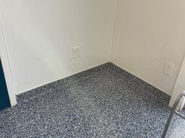 Empty corner of a room with white walls and speckled gray carpet, near a doorway and electrical outlets