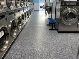Row of stainless steel washers and dryers in a laundromat with a blue bench on a speckled floor