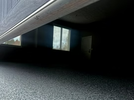 Dim room viewed from under a bed, with a window and closed door in the background.