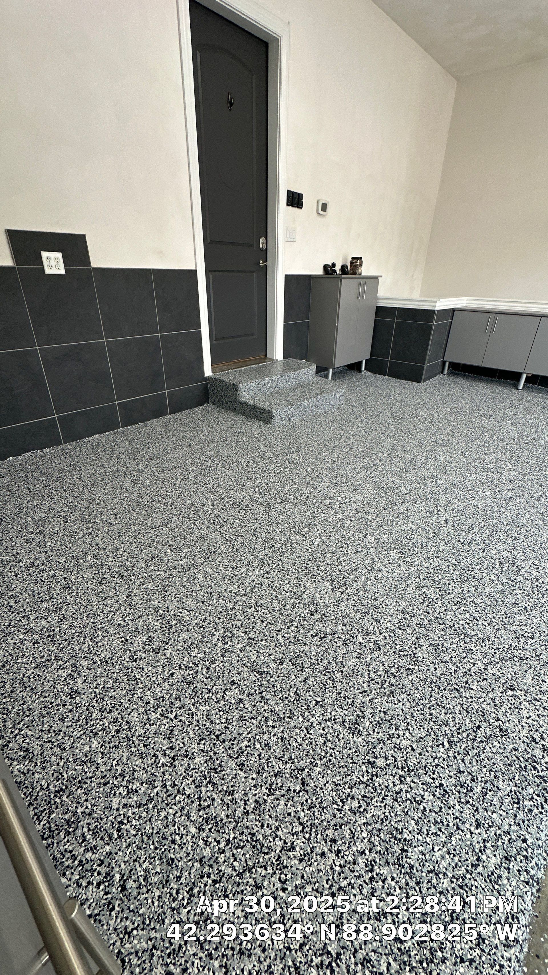 Empty indoor room with speckled floor, white walls, dark door, and a few cabinets along the right wall