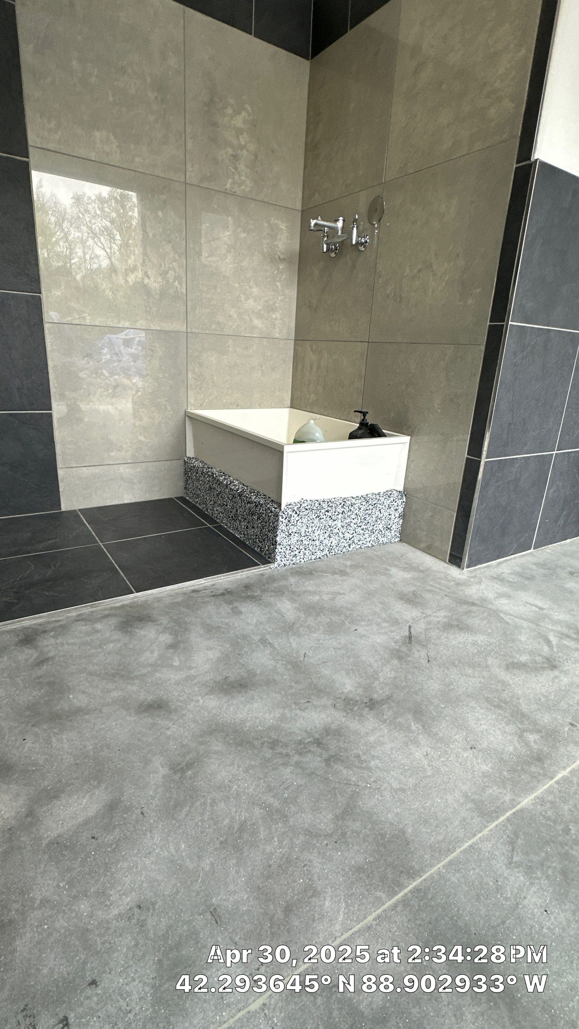 Small outdoor shower nook with beige tile walls, white sink bench, and gray floor tiles.