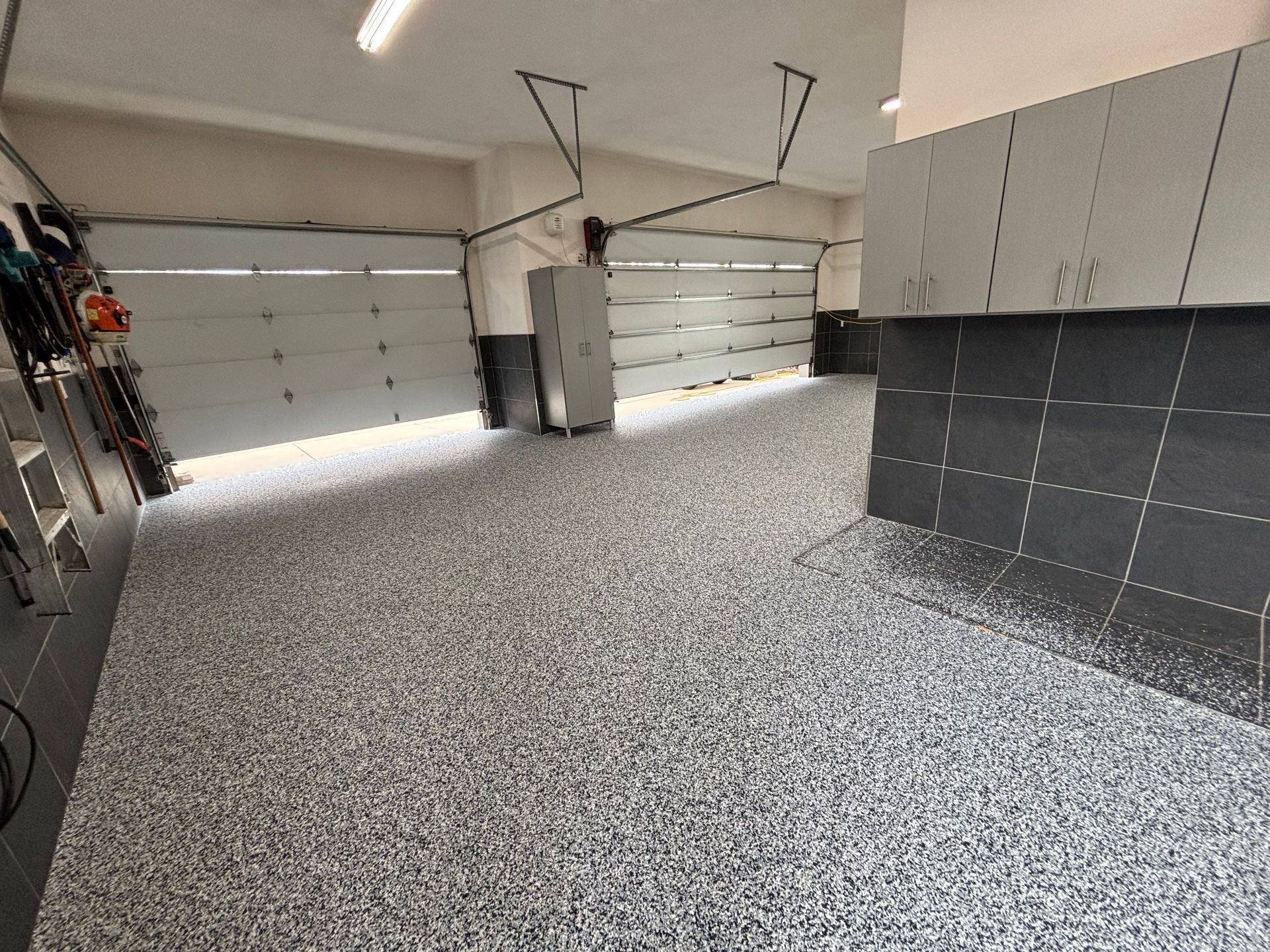 Empty garage with speckled floor, two white garage doors, and gray cabinets along the wall