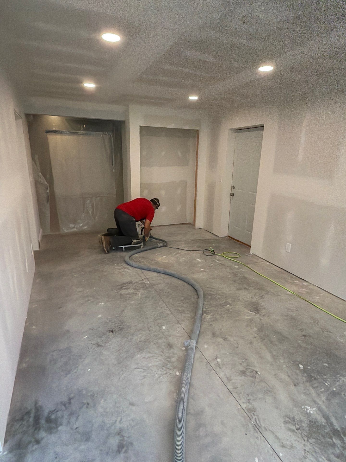 Worker using a floor grinder in a white room under renovation, with dust hoses and unfinished walls.