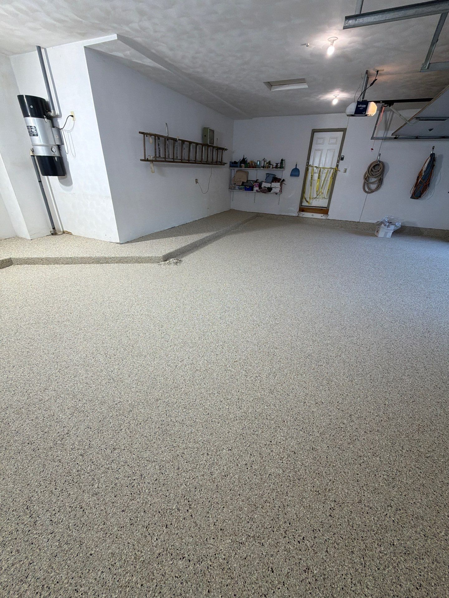 Empty garage with speckled floor, white walls, ceiling lights, and a ladder mounted on the left wall