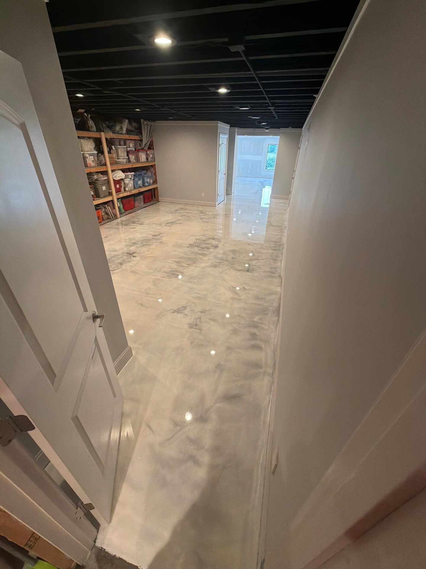 Narrow basement hallway with white walls, black ceiling, shelving, and plastic-covered floor leading to a lit doorway.