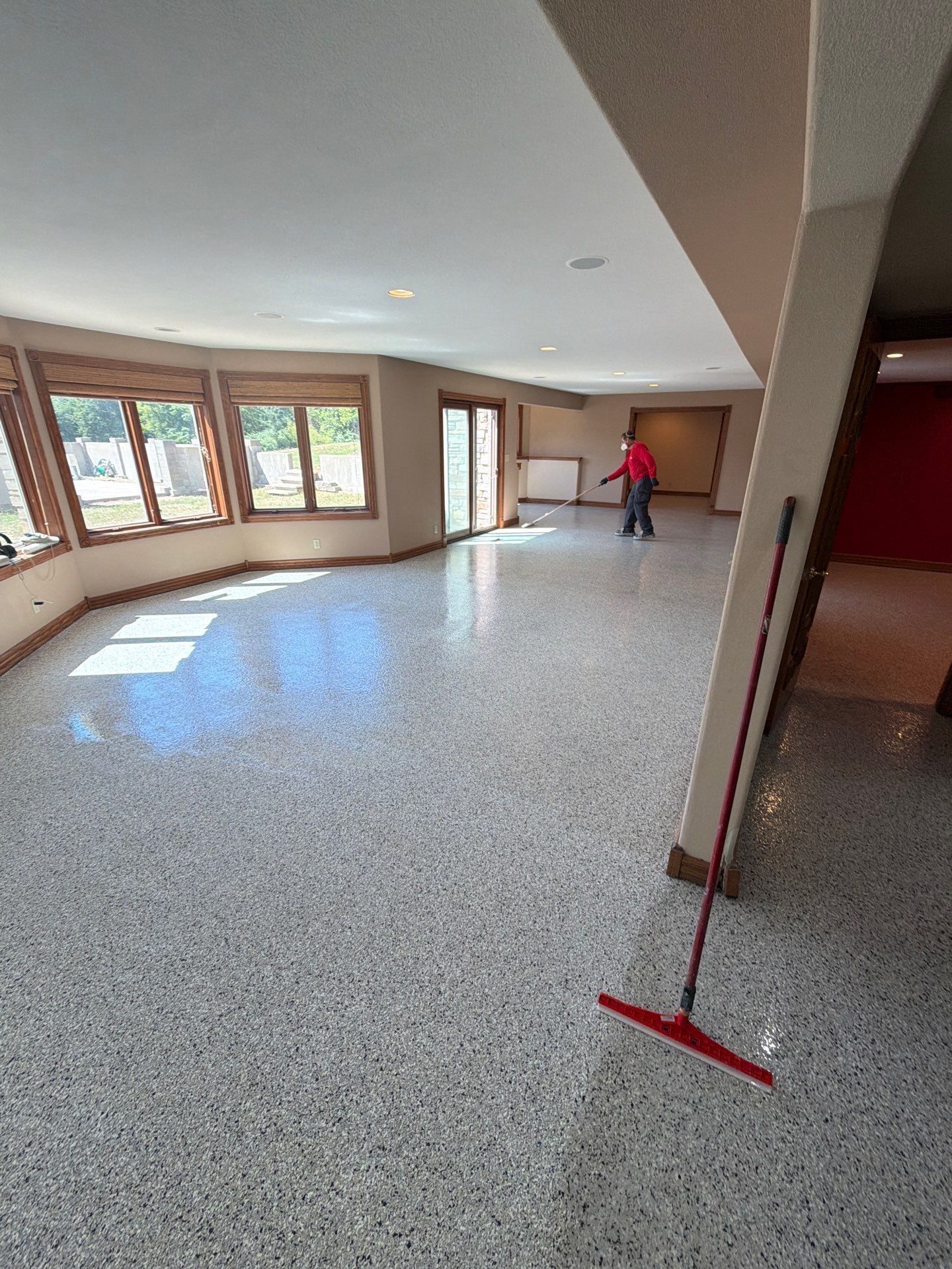 Empty room with speckled floor, large windows, and a person cleaning with a red mop.