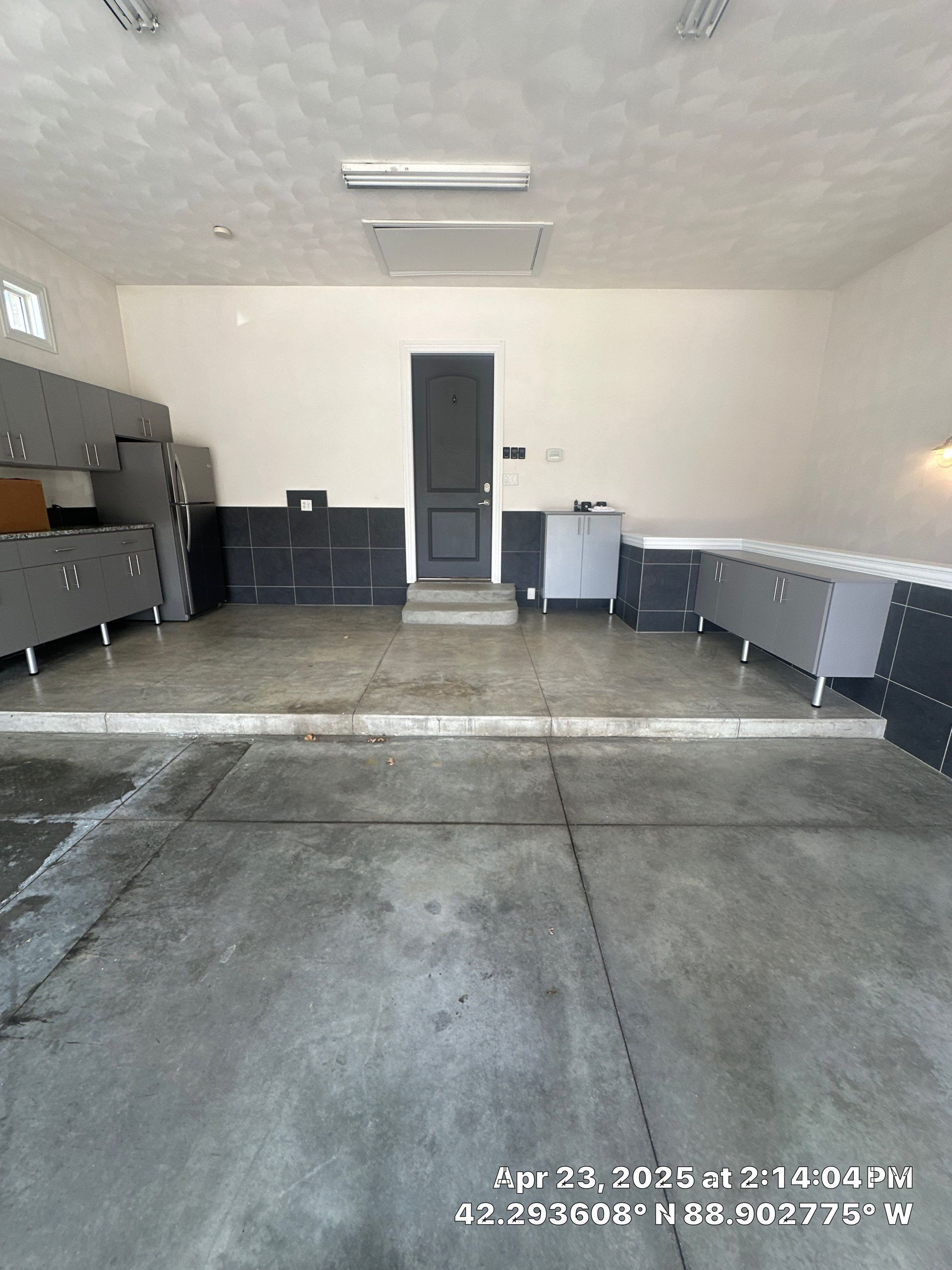 Empty garage with concrete floor, workbench on left, cabinets along walls, and a white door at the back.