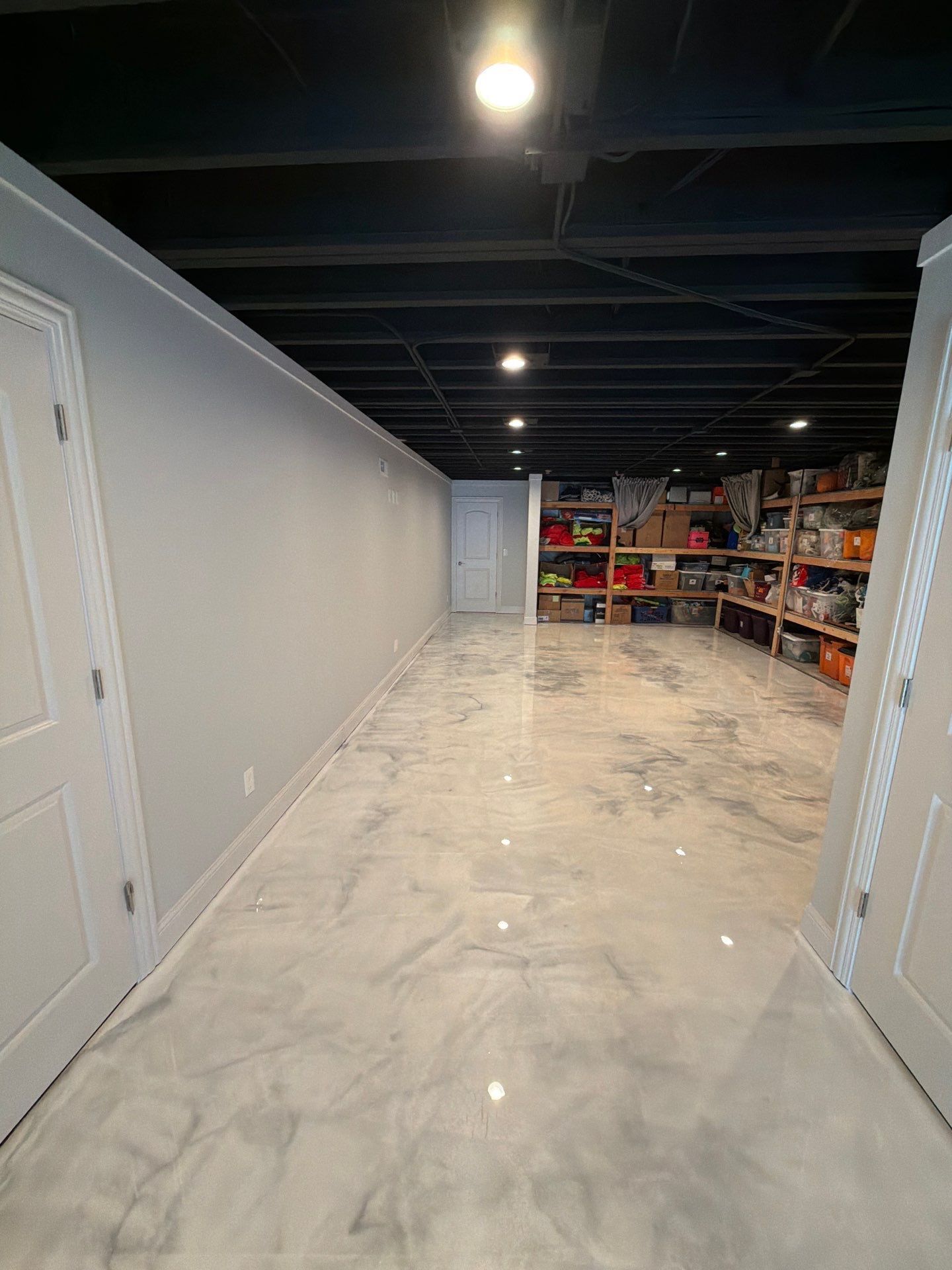 Long empty garage with gray walls, glossy concrete floor, black ceiling, and storage shelves at the back.