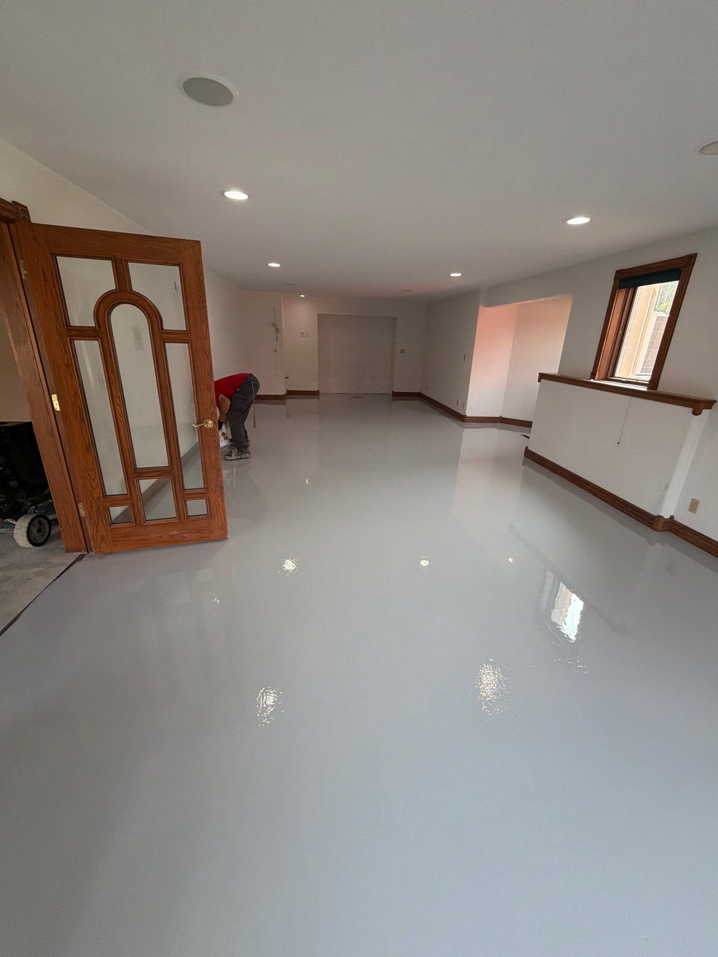 Bright empty room with glossy white floor, recessed lights, windows, and a wooden door panel at left.