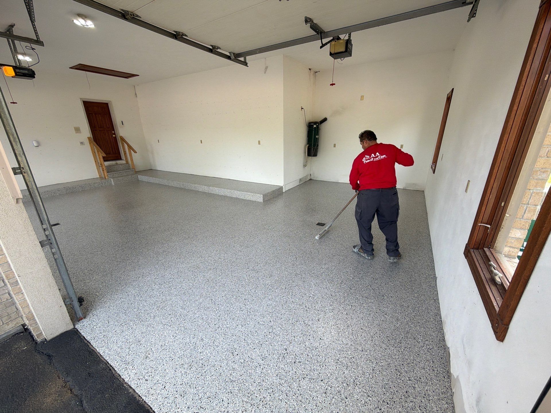 Worker in red shirt sweeping a large empty garage with speckled floor and white walls.