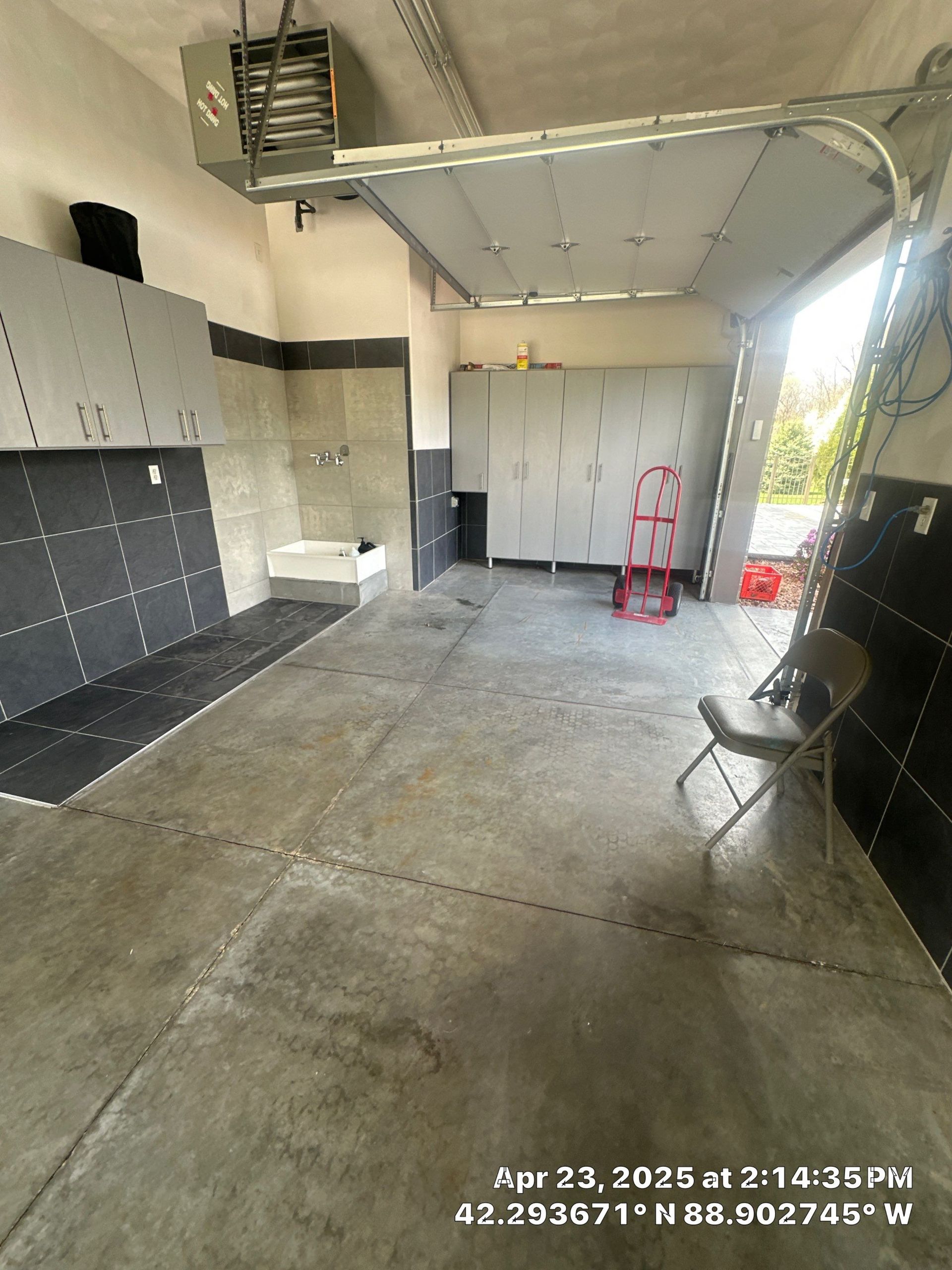 Empty industrial garage with concrete floor, cabinets, and folding chair near a bright open doorway.