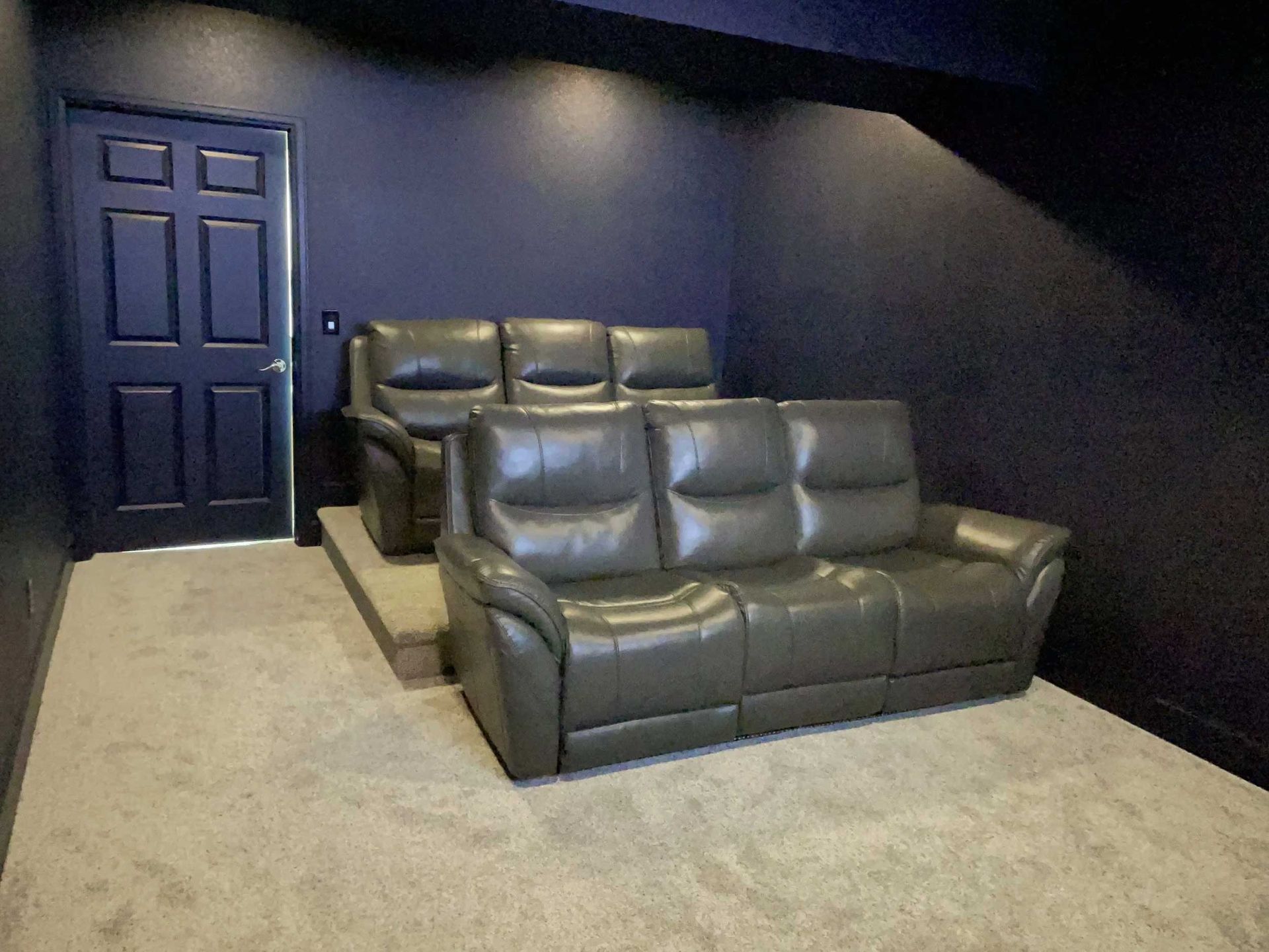 A dark-walled home theater with gray leather couches on a raised platform. A door is on the left.