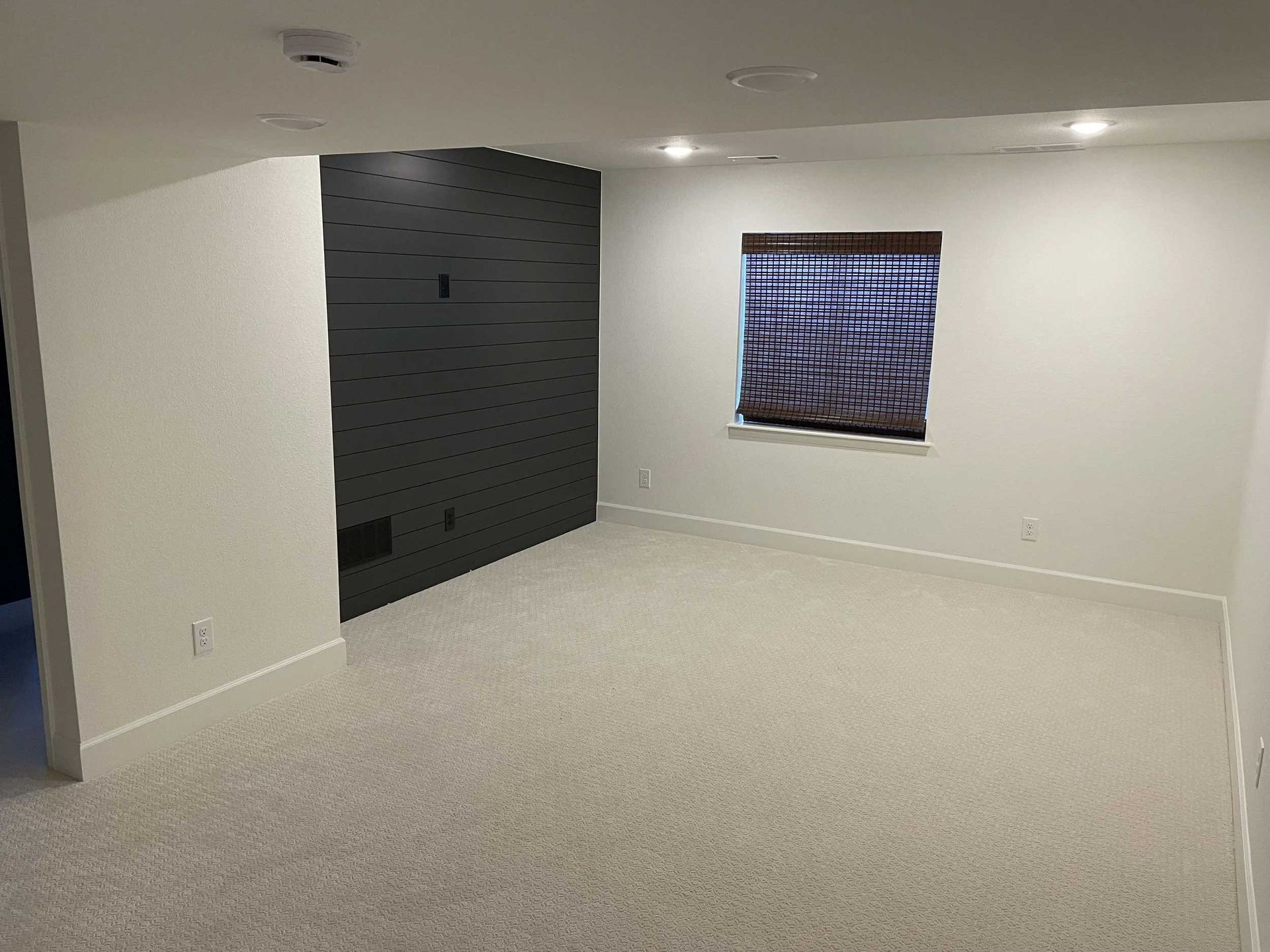 Empty room with light carpet, dark gray accent wall, small window with blinds, and recessed lighting.