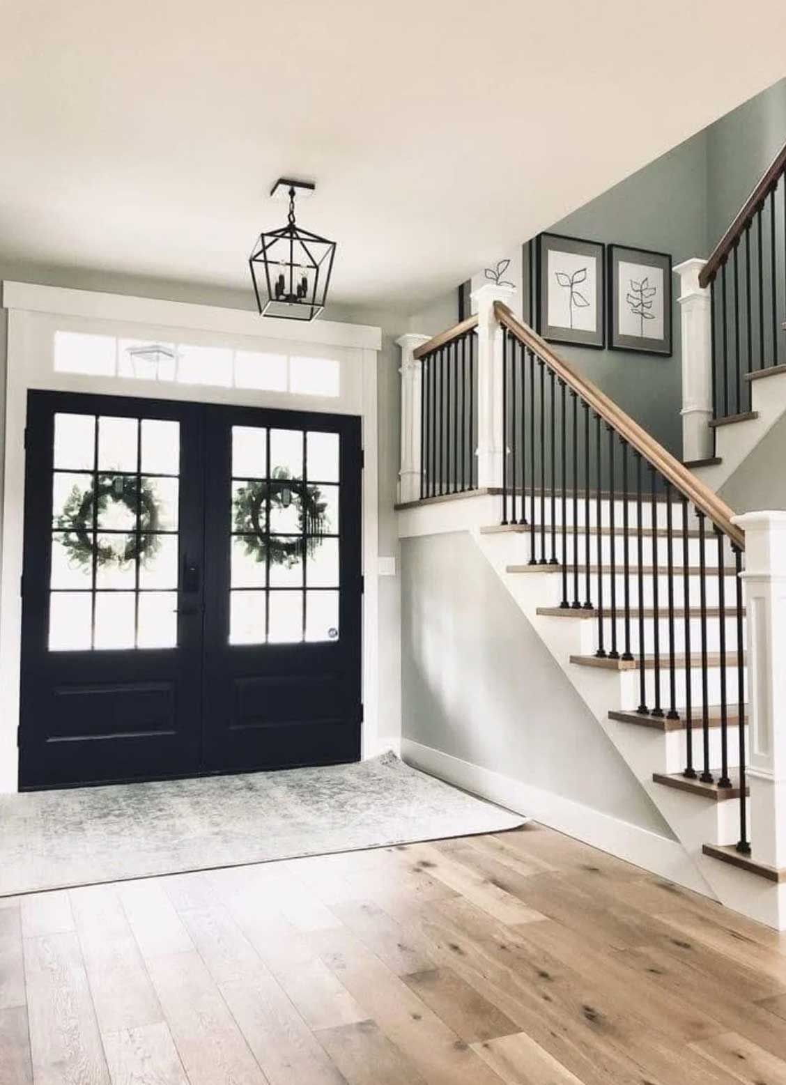 Entryway with black double doors, staircase, and neutral colors. Wreaths hang on the doors, and artwork hangs on the gray wall.