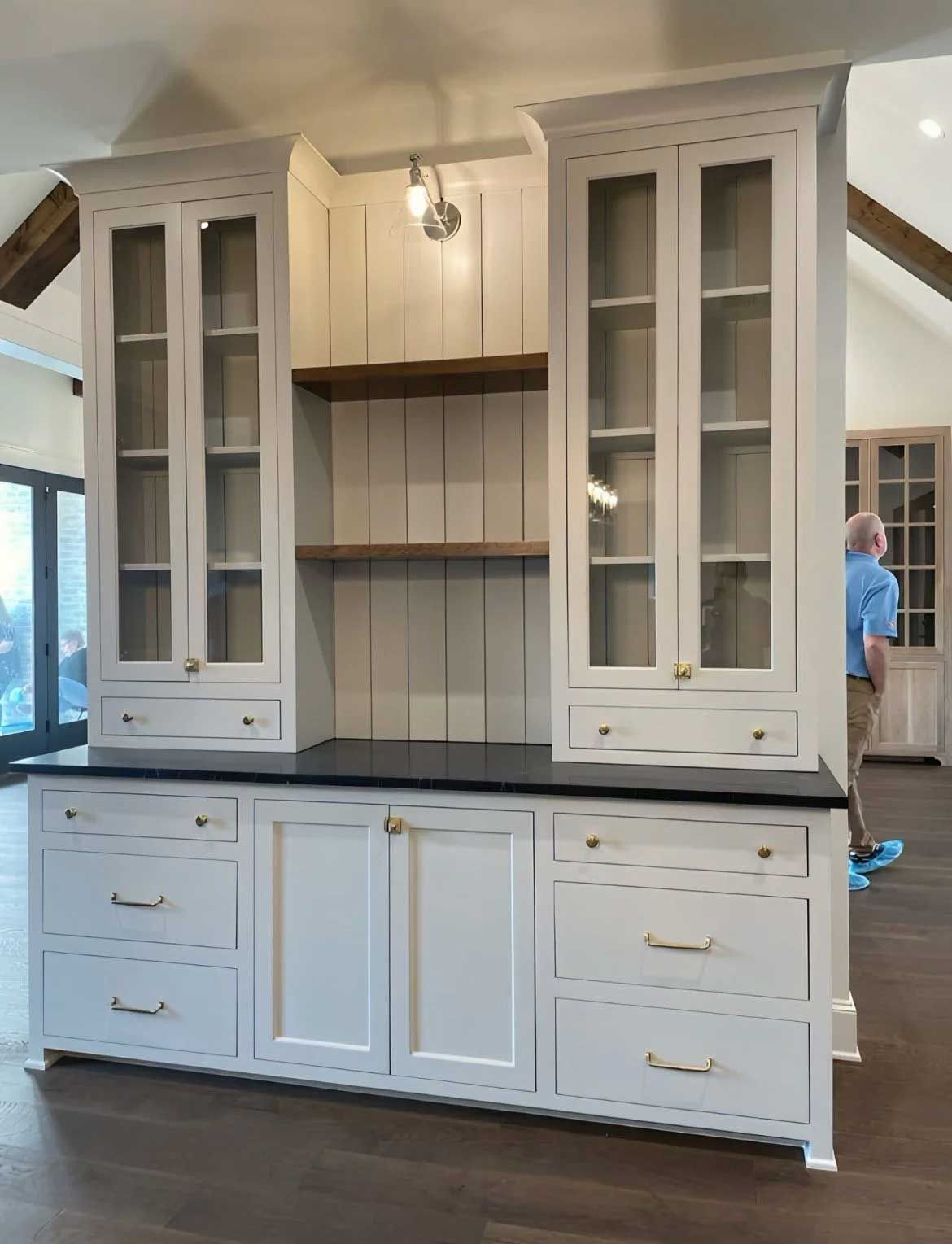 White kitchen hutch with glass-door cabinets, drawers, and black countertop. A person stands nearby.