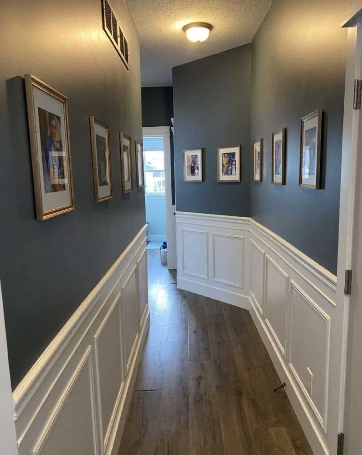 A long hallway with dark blue walls, white wainscoting, framed pictures, and a wood-look floor.