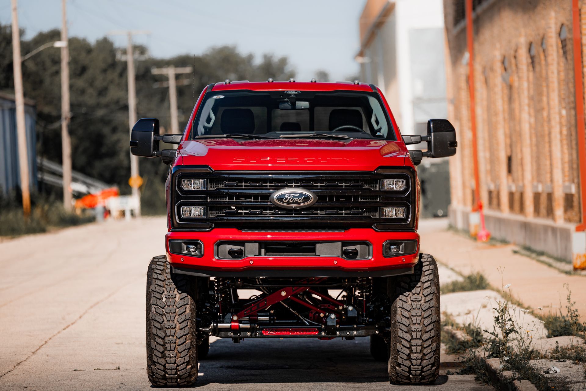 Red lifted Ford truck centered on a road, black grill and mirrors, large tires.