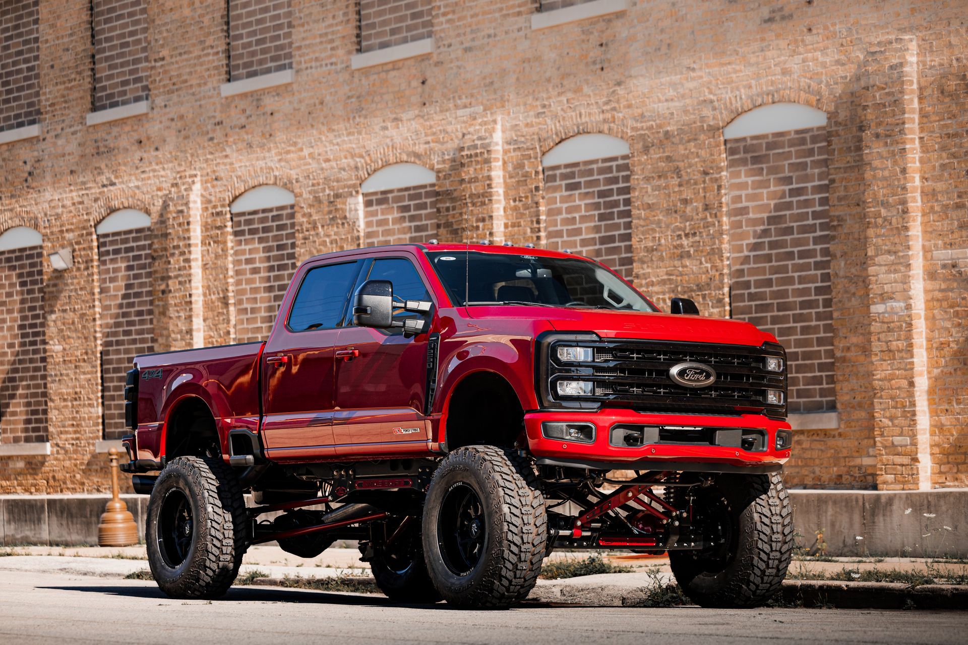 Red lifted Ford truck with black tires parked in front of a brick building.