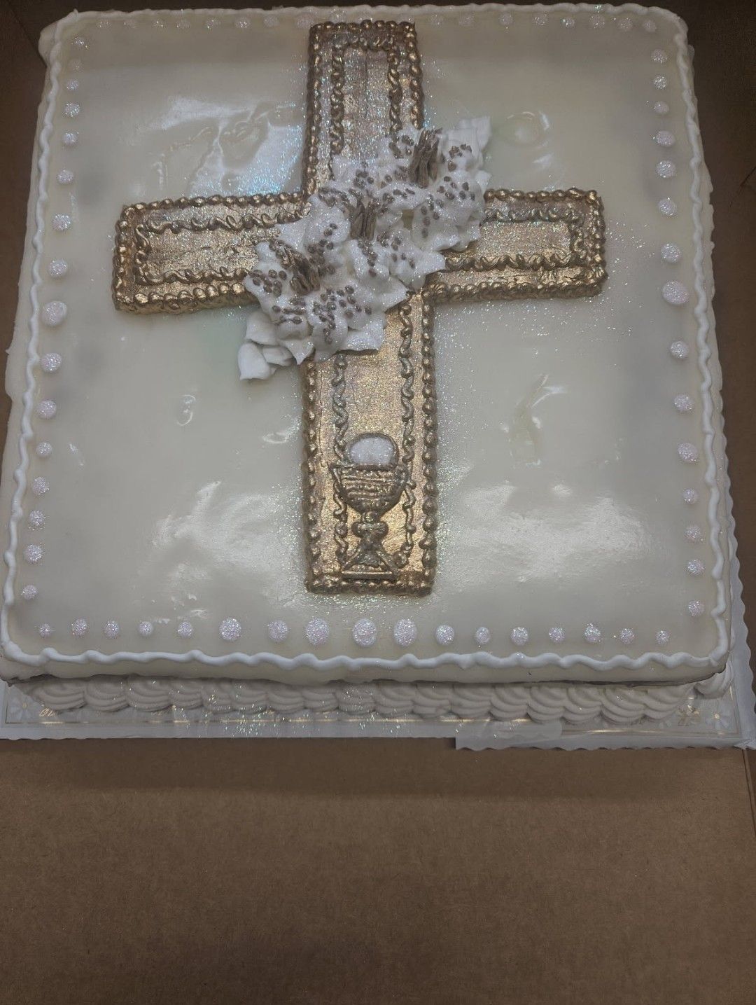 Square cake with white frosting and a gold cross decorated with white flowers and pearl border