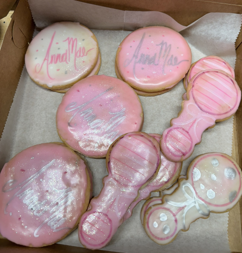 Pink and white decorated cookies in a box, including round and bottle-shaped treats with “Mia” written on one.