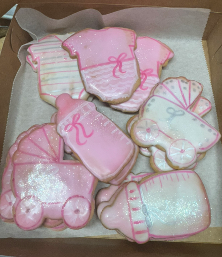 Pink and white baby-themed decorated cookies in a box, with onesies, booties, and a carriage