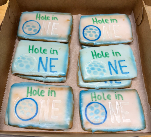 Six frosted cookies in a cardboard box, each decorated with “Hole in ONE” golf-themed icing.