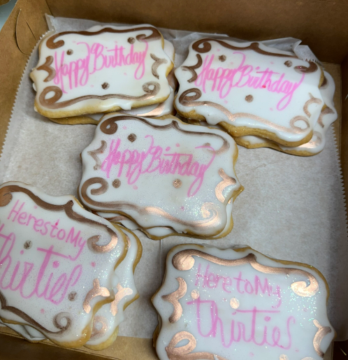 Decorated birthday cookies with pink icing and gold swirls in a box, some reading “Happy Birthday” and “Auntie.”