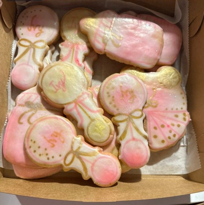 Pink and gold decorated cookies in a bakery box, shaped like baby rattles and onesies.