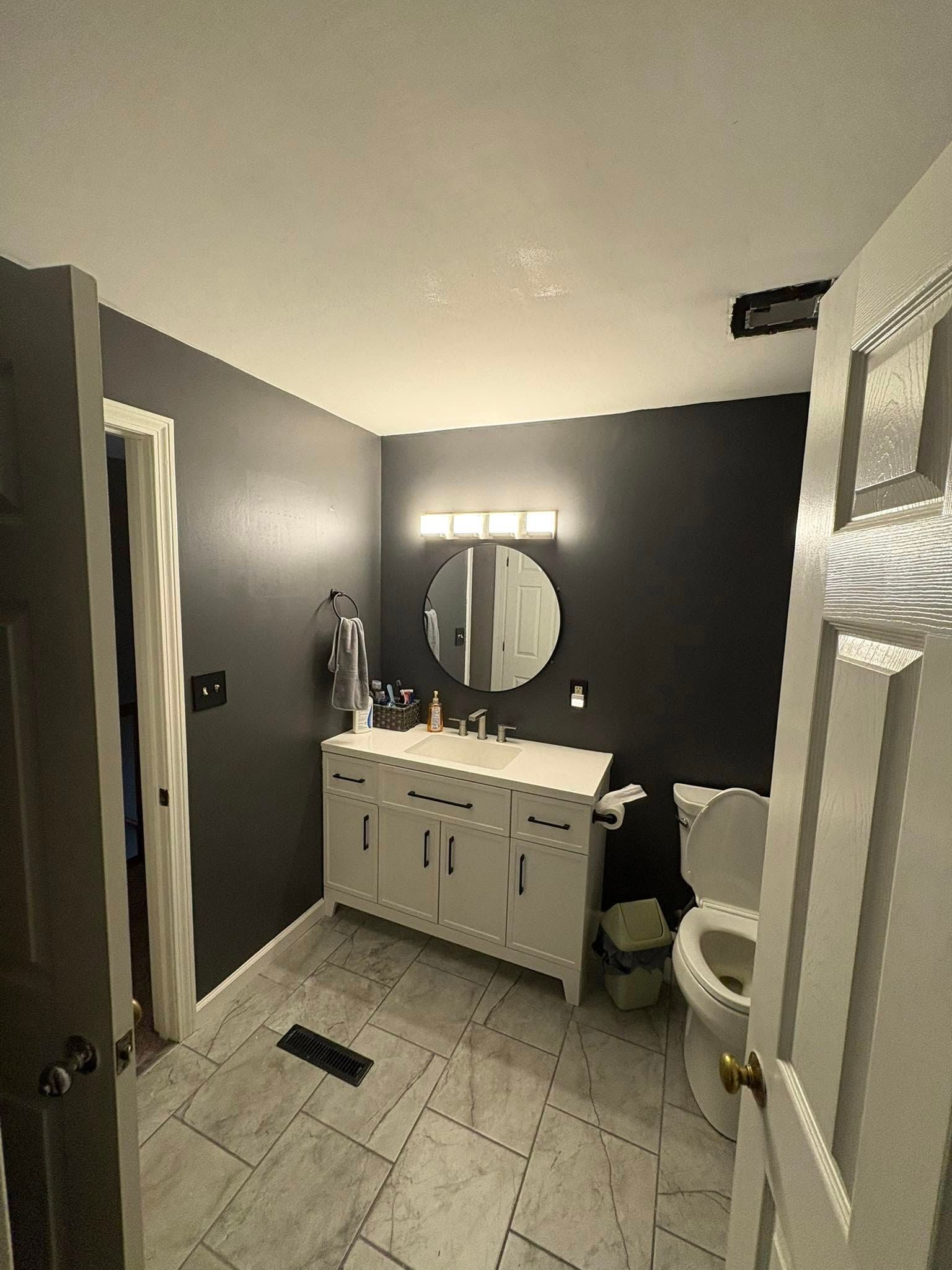 A dark gray bathroom with a white vanity, round mirror, and open white door.