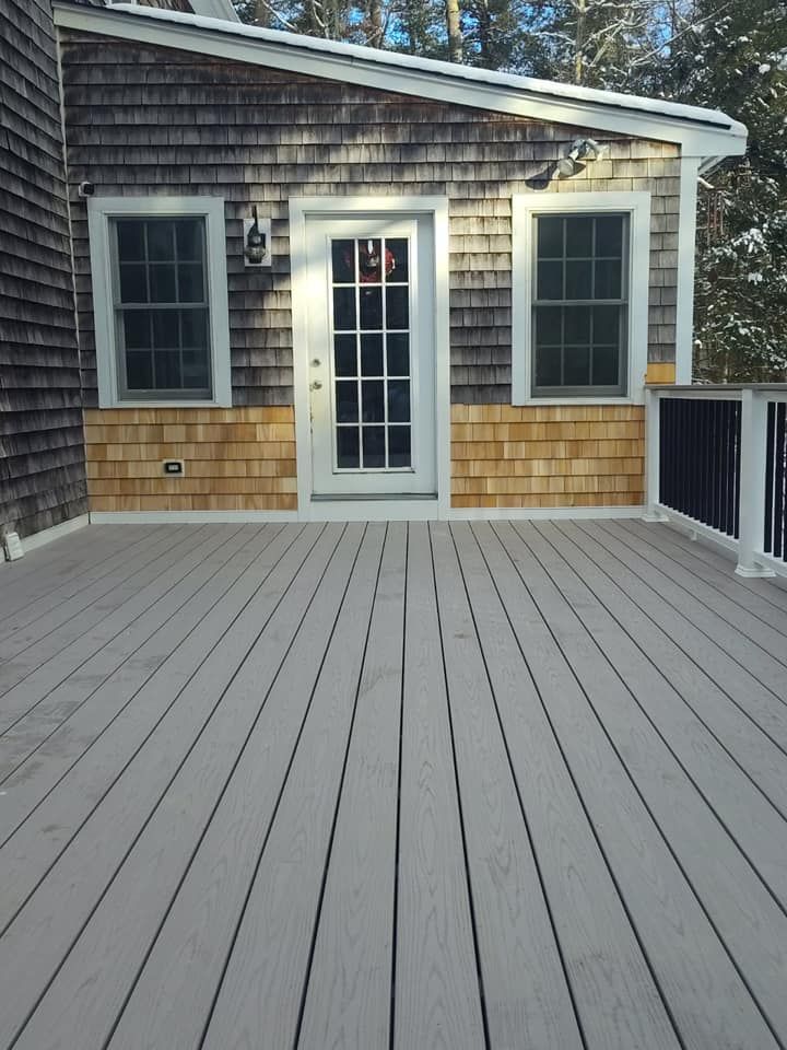 Deck with gray composite decking, a white door, and cedar-shingled walls. Black railing on the right.