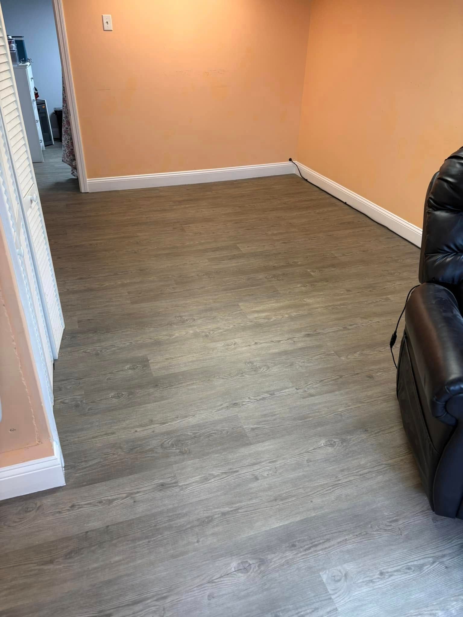 Room with gray wood-look flooring, tan walls, white trim, and a black recliner. A doorway is on the left.