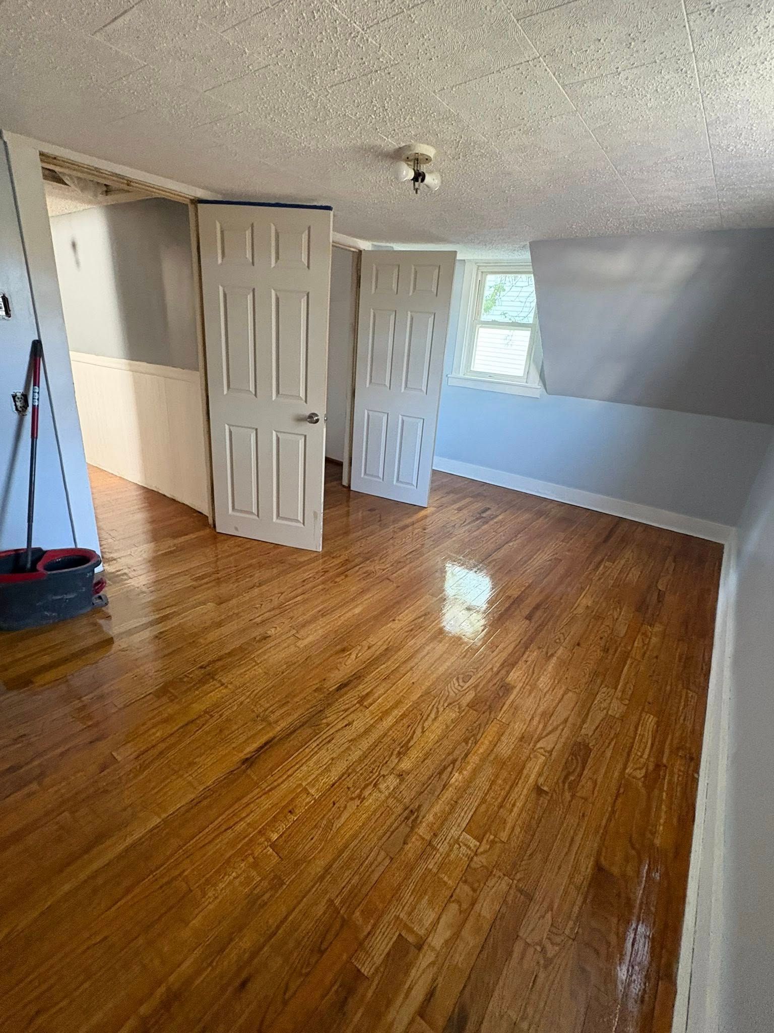 Wooden-floored room with two white doors, small window, white and gray walls, and cleaning supplies.