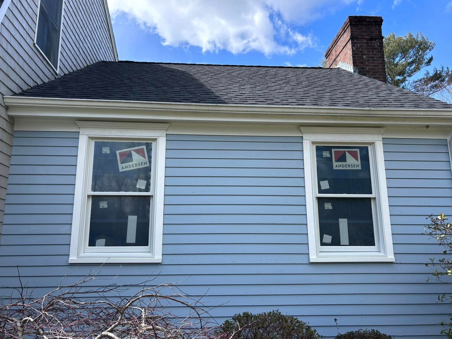 Blue house side with two white-framed windows, gray roof, and a brick chimney under a partly cloudy sky.