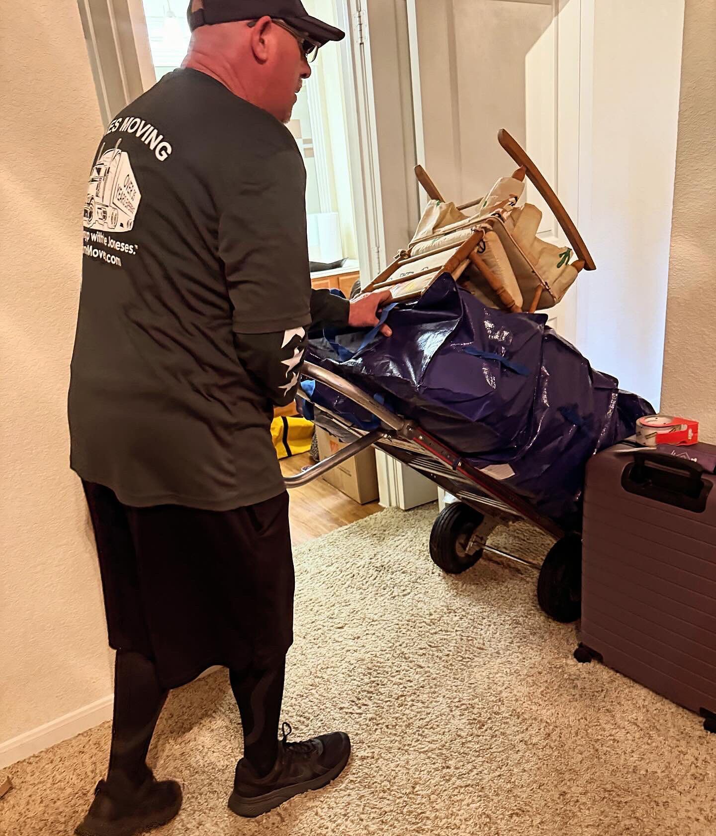Man in black shirt and cap moving items on a hand truck through a doorway; indoor, moving day.