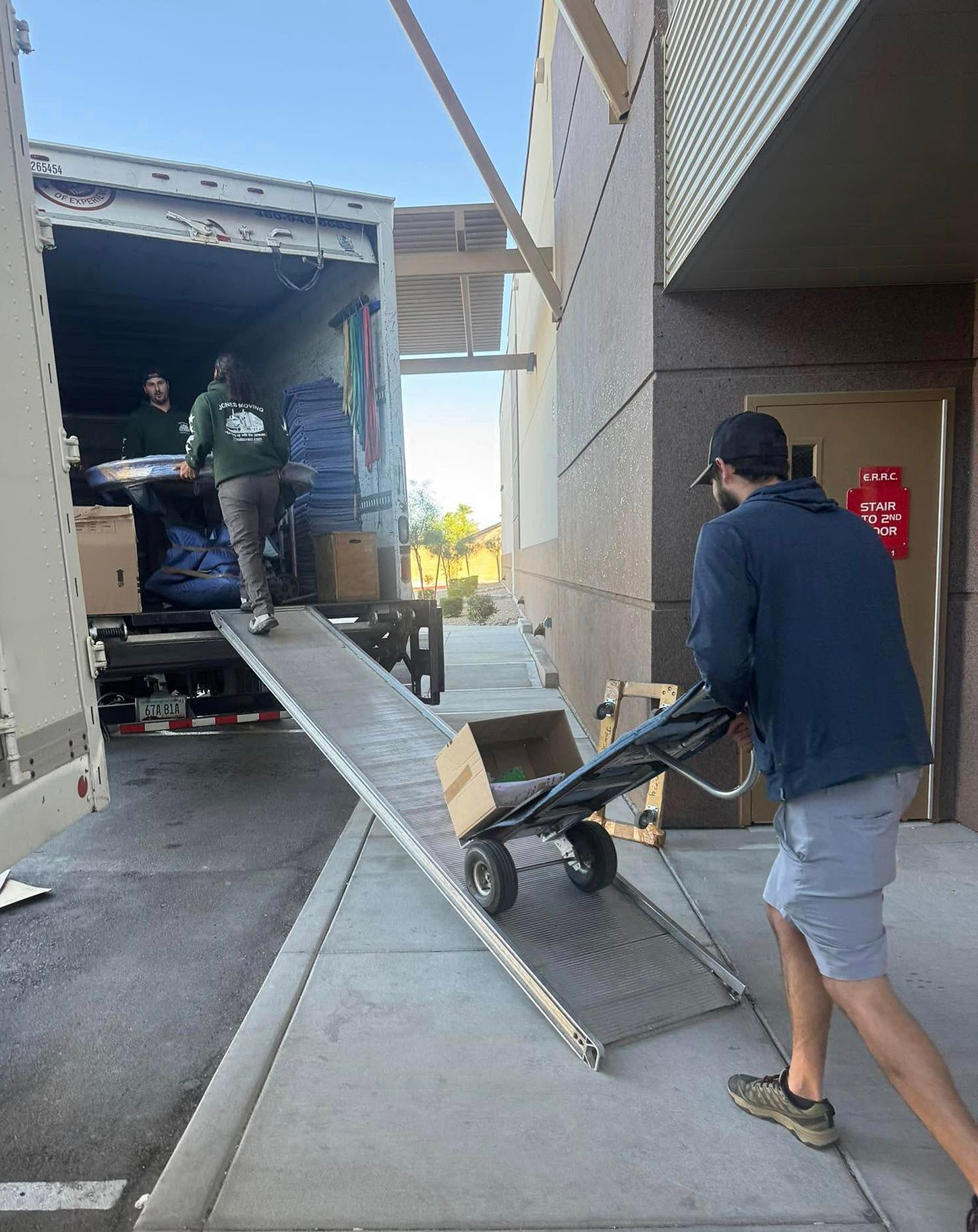 People moving boxes from a truck, using a ramp and hand truck, at a building with a concrete walkway.
