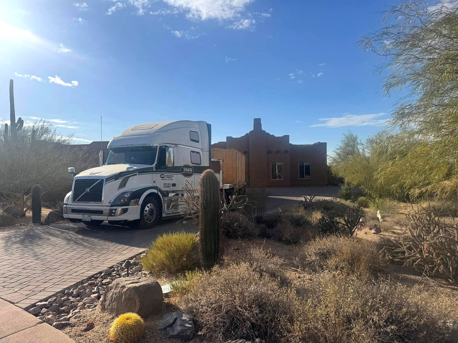 A semi-truck parked in front of a brown adobe house in a desert setting under a blue sky.