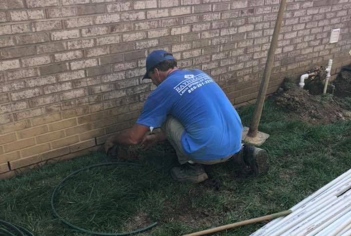 A man in a blue shirt is kneeling in front of a brick wall.
