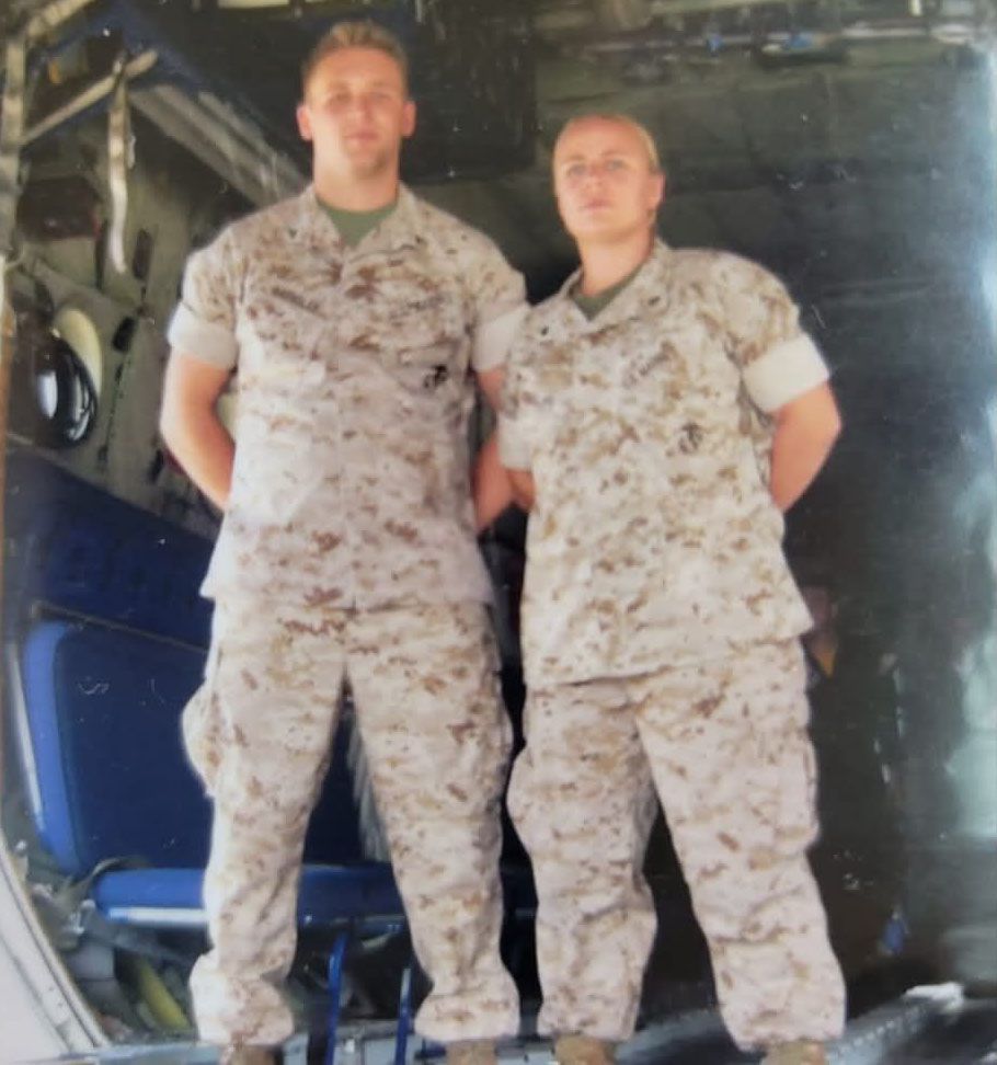 Two individuals in military desert camouflage uniforms standing side-by-side inside the cargo bay of an aircraft.