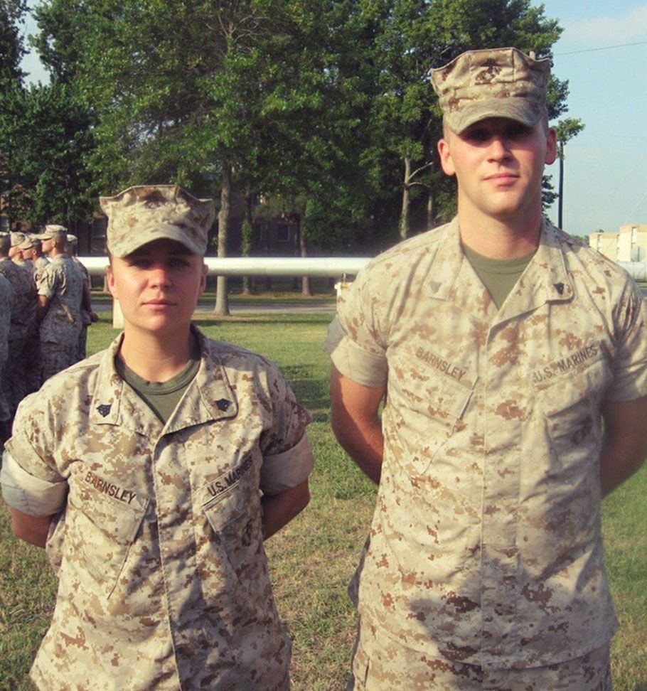 Two Marines in desert camouflage uniforms standing at attention on a grassy field with trees and pipes in the background.