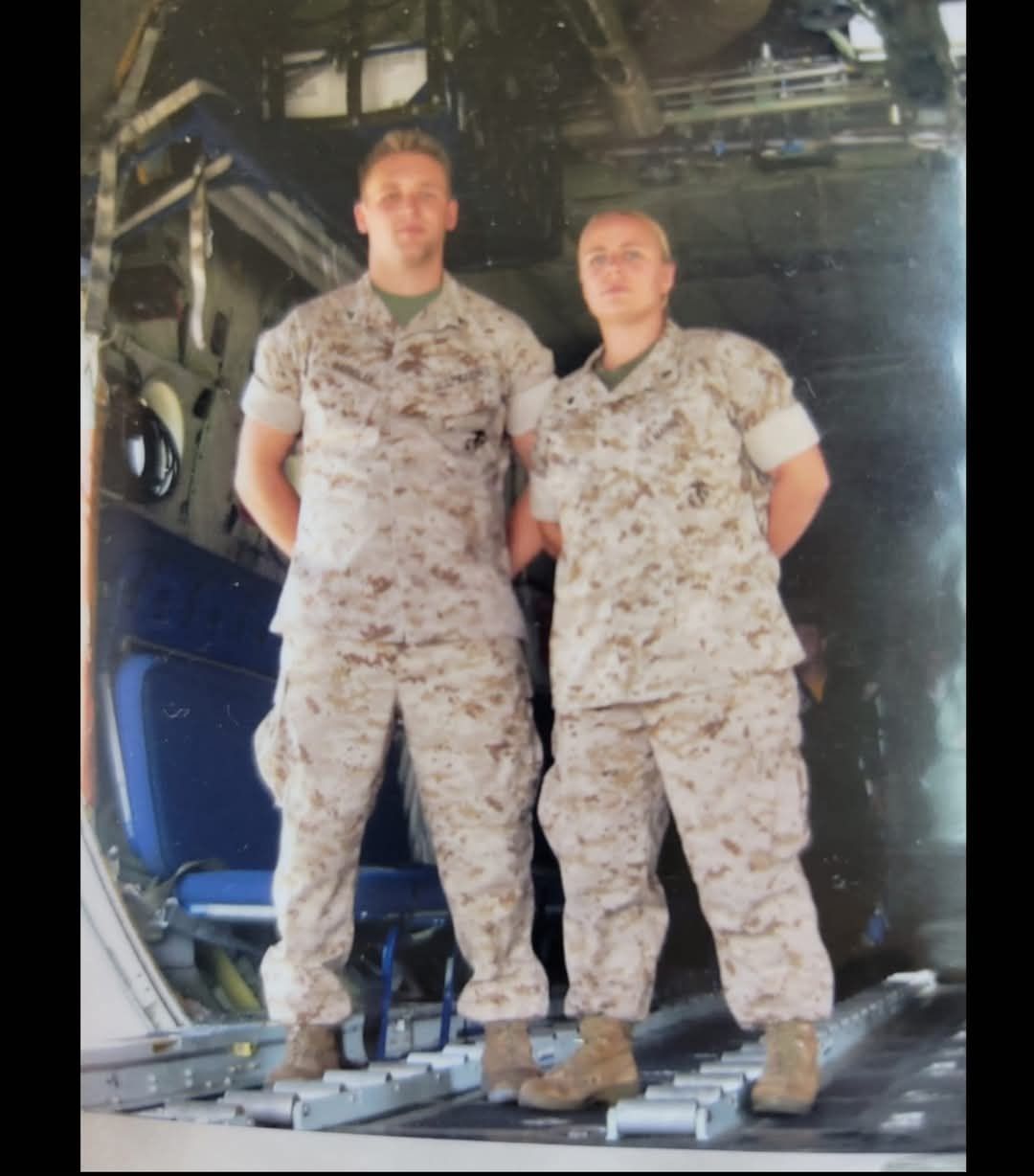 Two individuals in military desert camouflage uniforms standing side-by-side inside the cargo bay of an aircraft.
