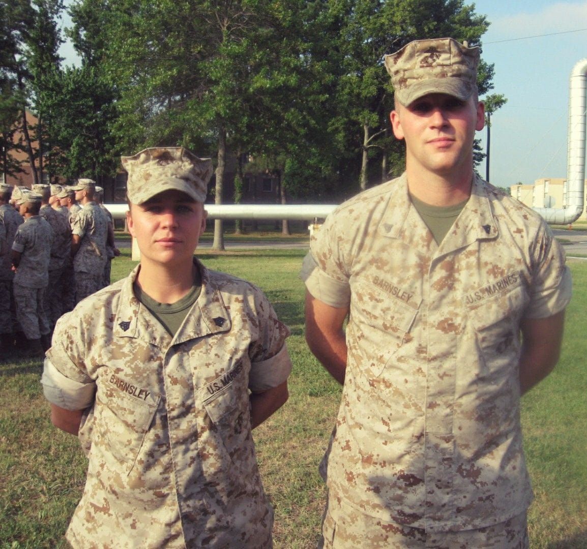 Two Marines in desert camouflage uniforms standing at attention on a grassy field with trees and pipes in the background.