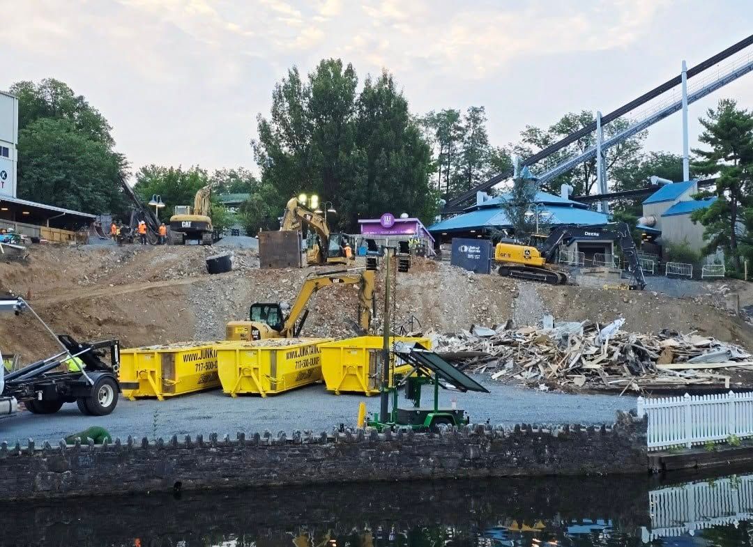 Construction site with yellow dumpsters and excavators; a rollercoaster in the background.