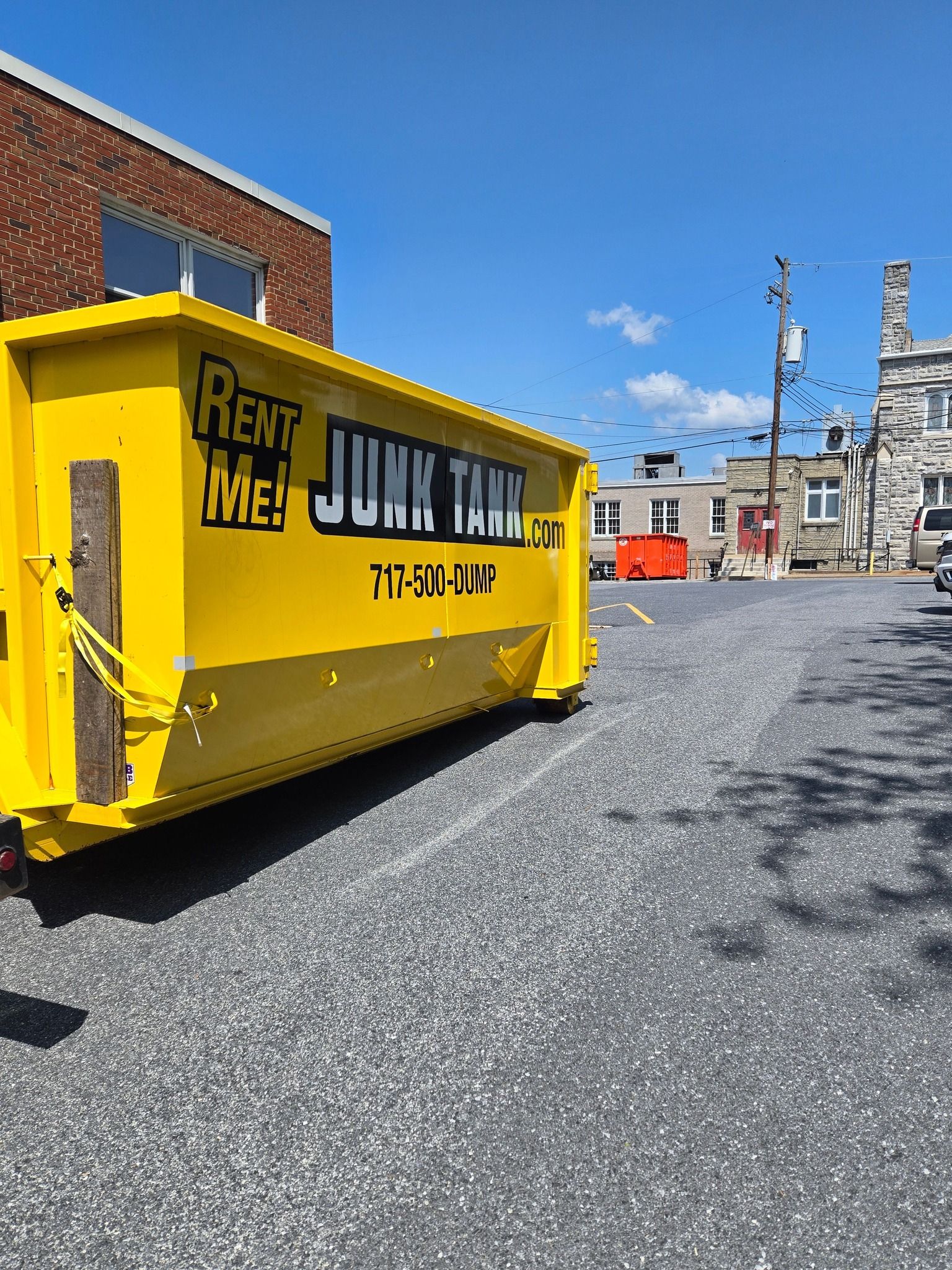 Yellow junk removal dumpster on a paved lot, with a brick building in the background on a sunny day.