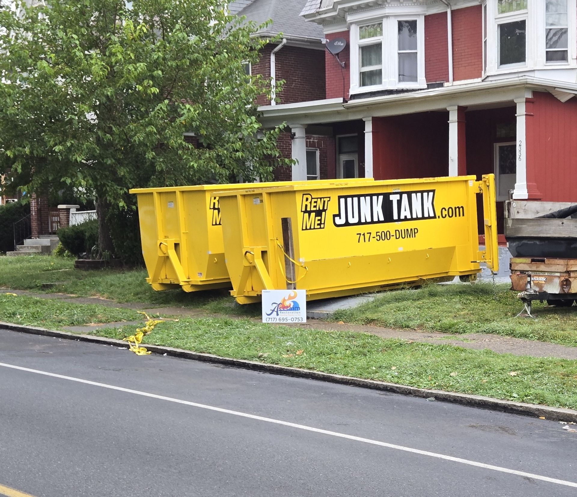 Yellow dumpster on residential lawn near a red house. Sign in front.