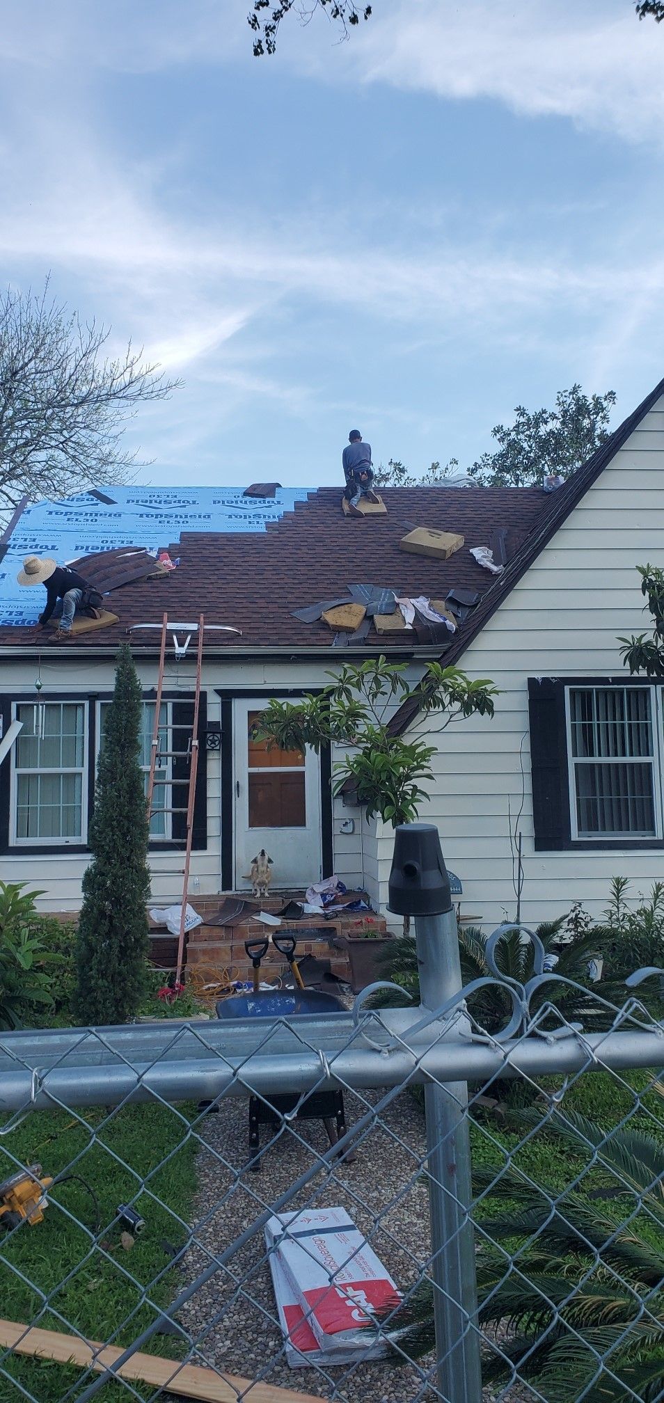 A man is working on the roof of a house behind a chain link fence.
