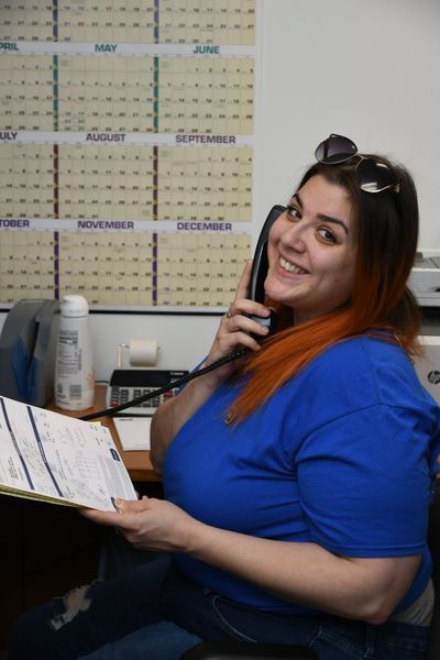 Woman in blue shirt, holding phone and papers, smiling at the camera in an office setting with a calendar.