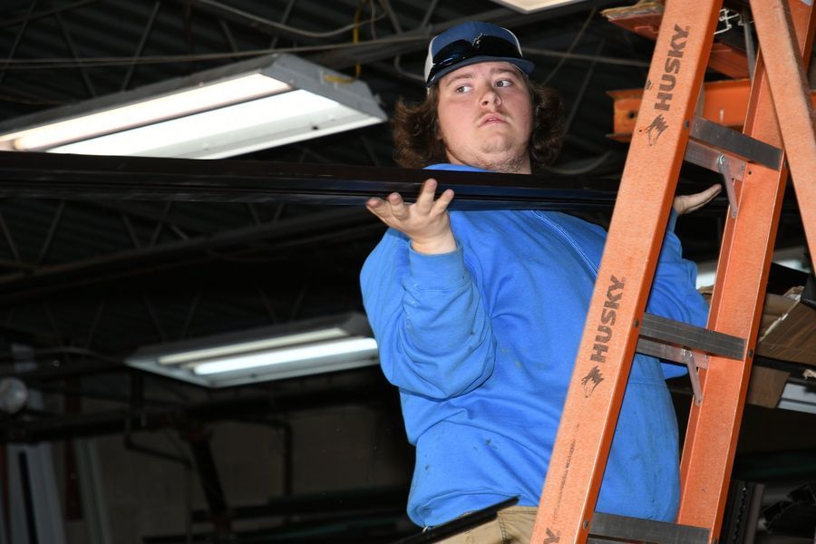 Man on ladder, holding a long, dark object in a workshop. Wearing blue shirt, cap, and safety glasses.